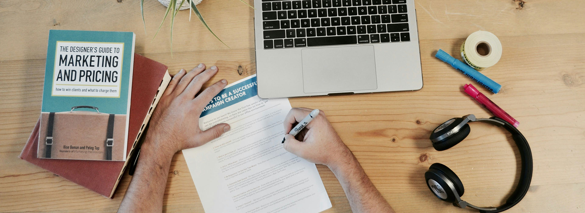 person writing at a desk with a book, laptop, headphones