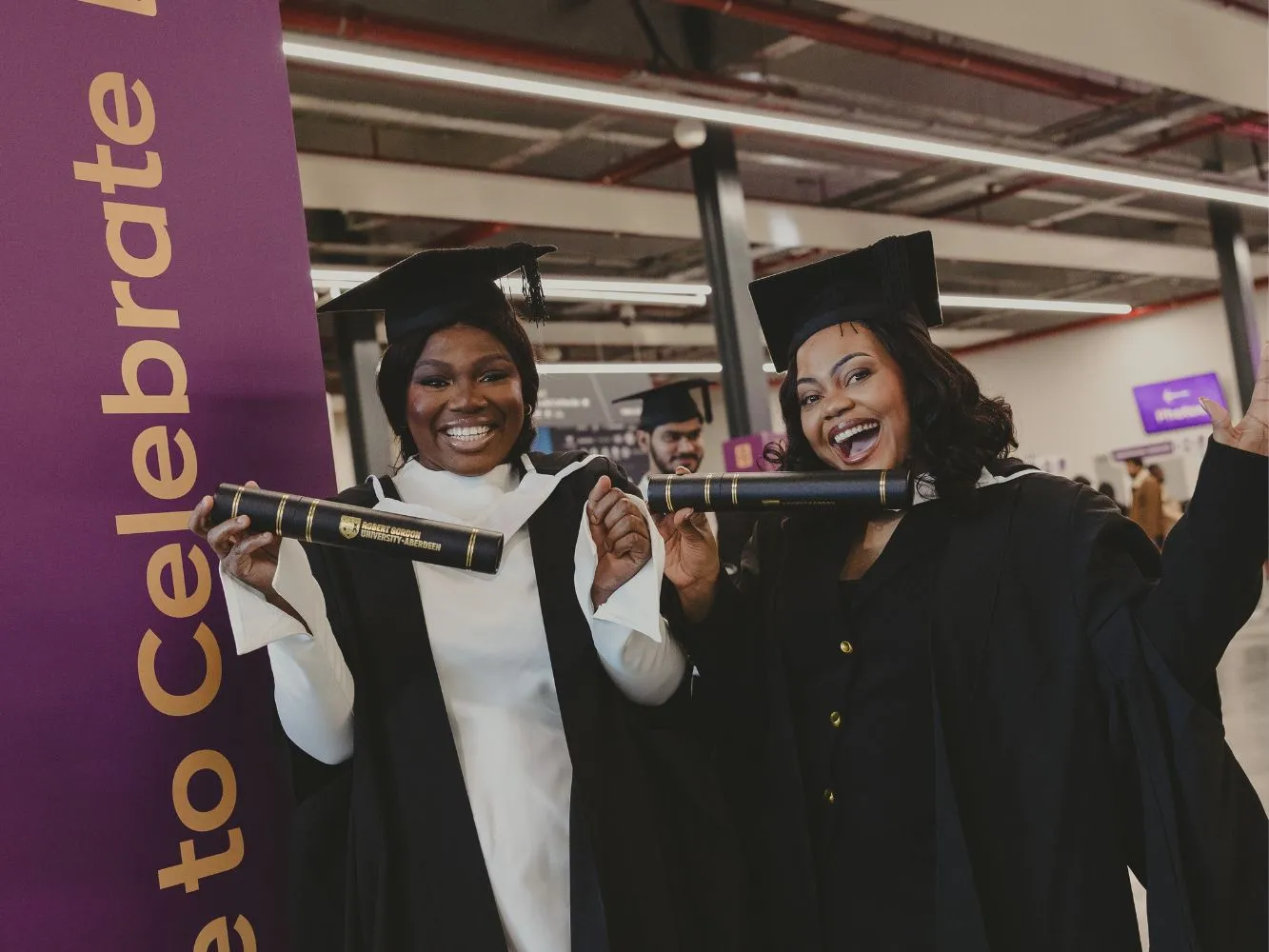 Two RGU graduates wearing caps and holding scrolls