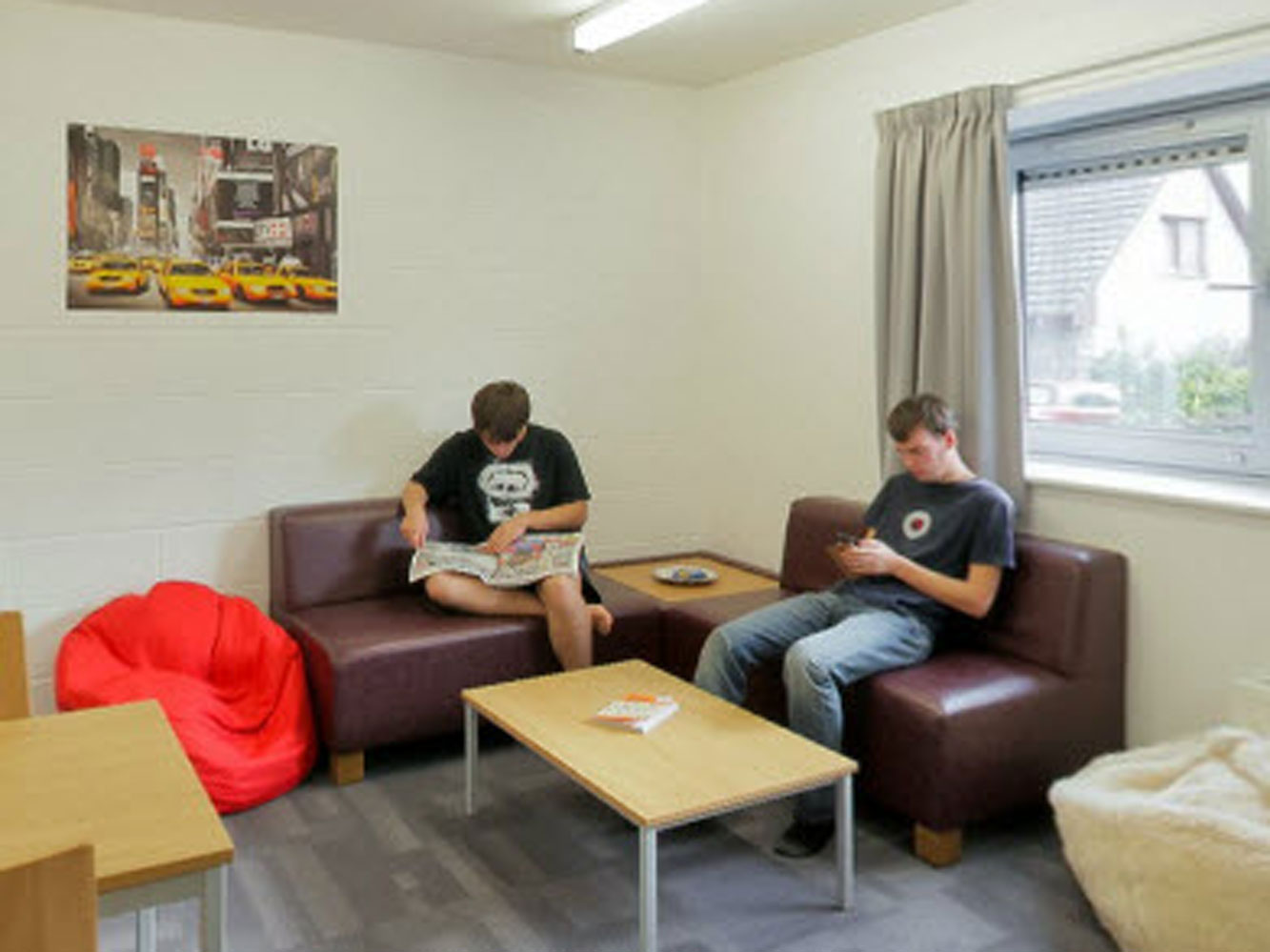 two students sitting in the lounge area, one reading from a newspaper and one using their phone