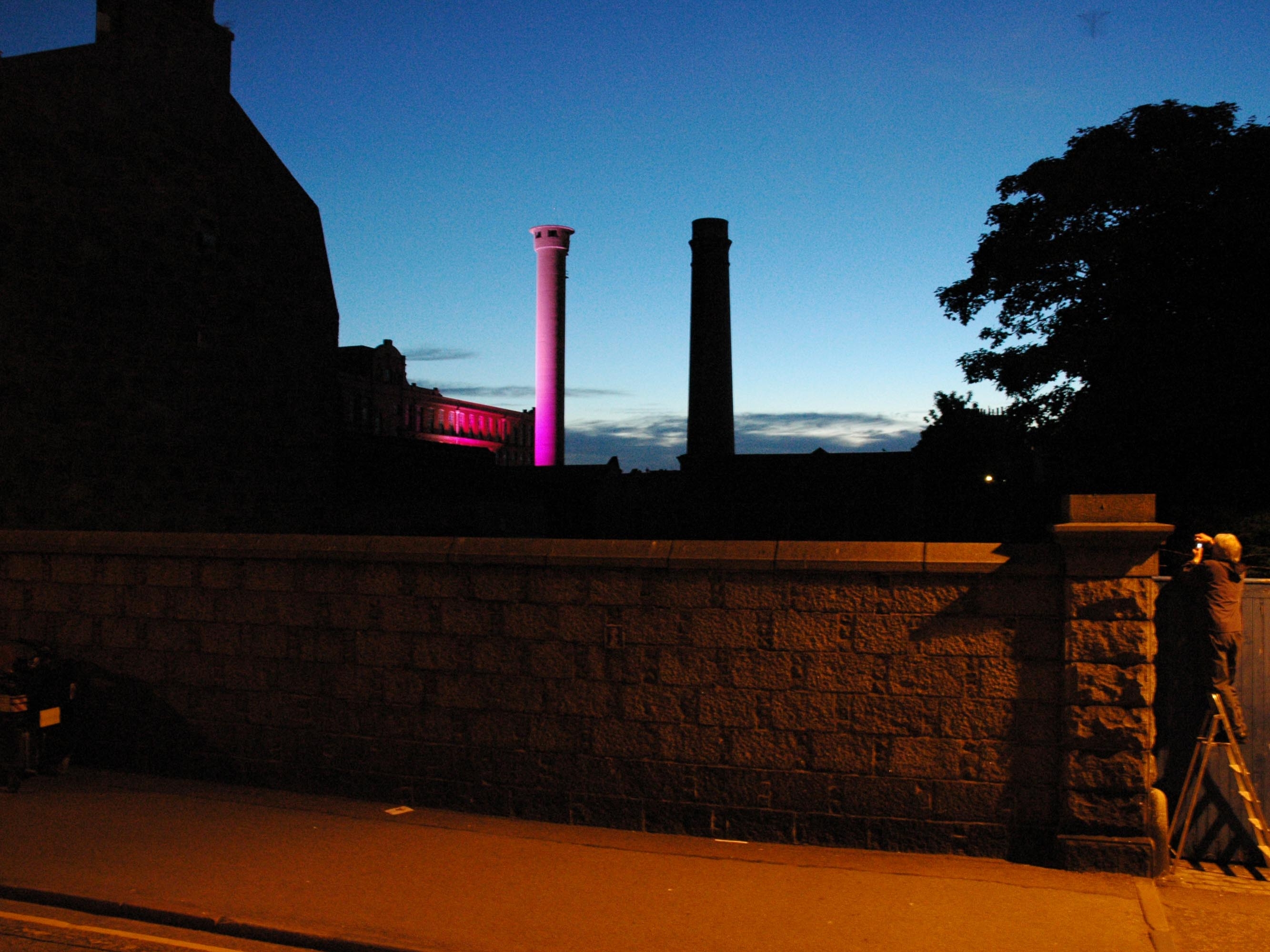 two factory towers seen over a granite wall, one is in dark shadow and the other is cast in a pink light