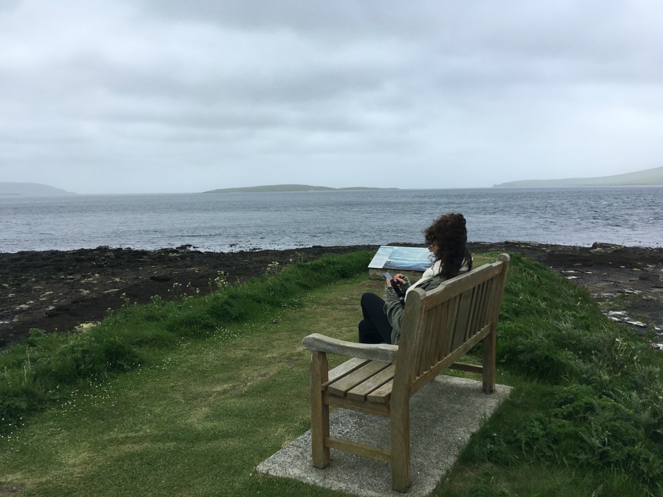 Person sitting on bench using phone with seascape in background