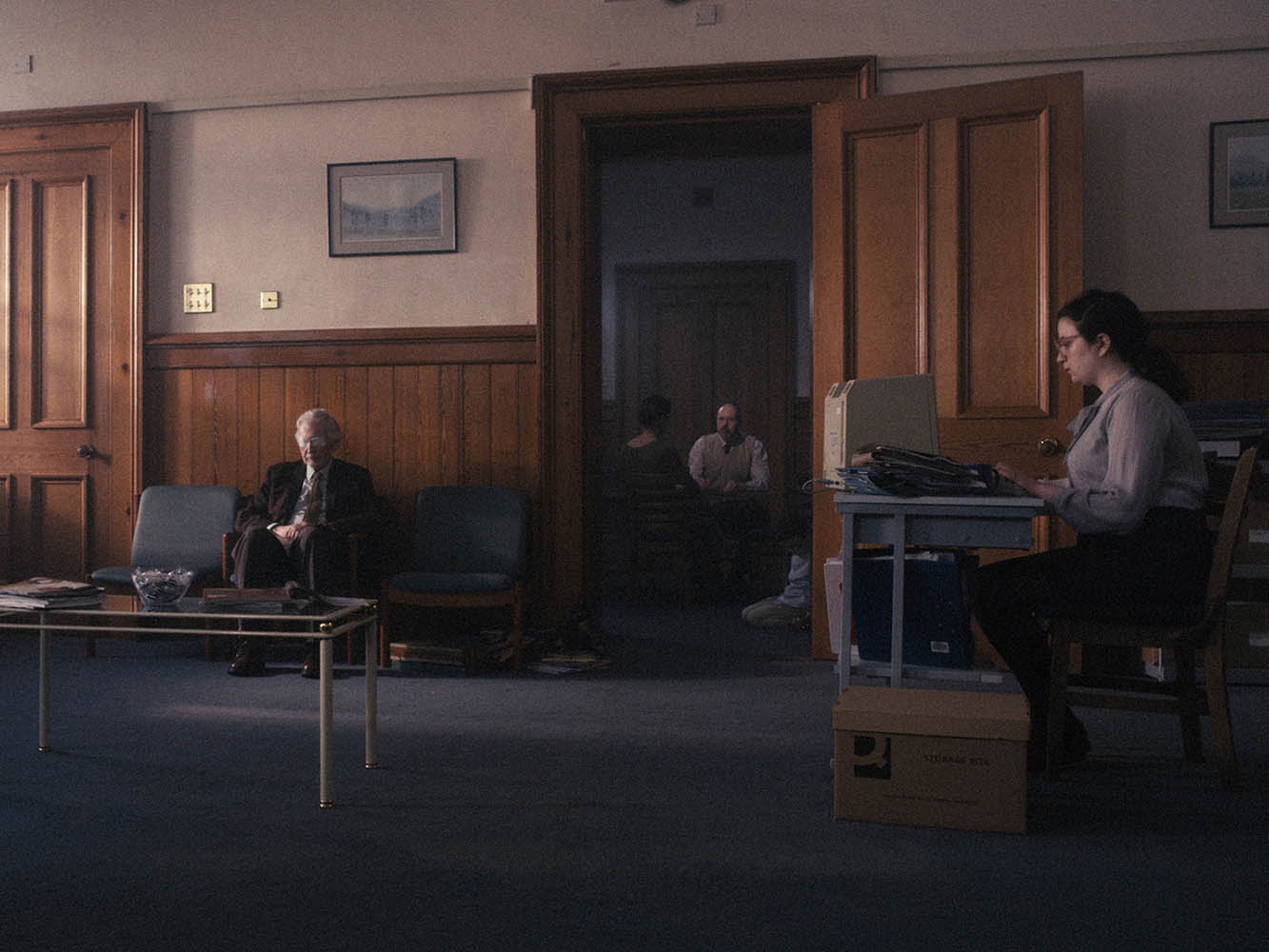 Reception room with woman sat at desk with a pile of files, older man waiting on a chair, through a door there is a man having a conversation with another person while a child kneels on the floor