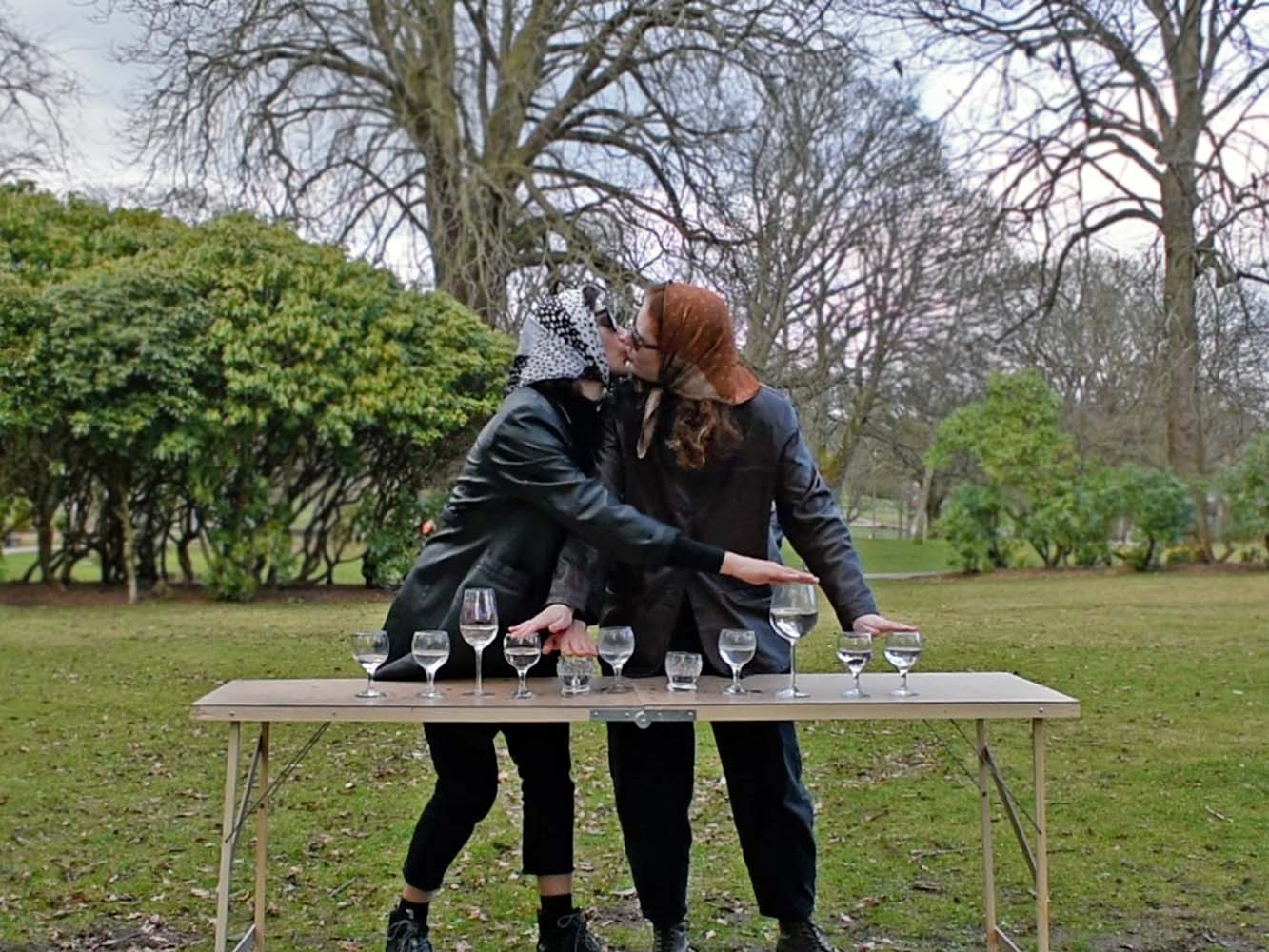 Two women wearing headscarves kiss in a park, touching wine glasses filled with water set on a table 
