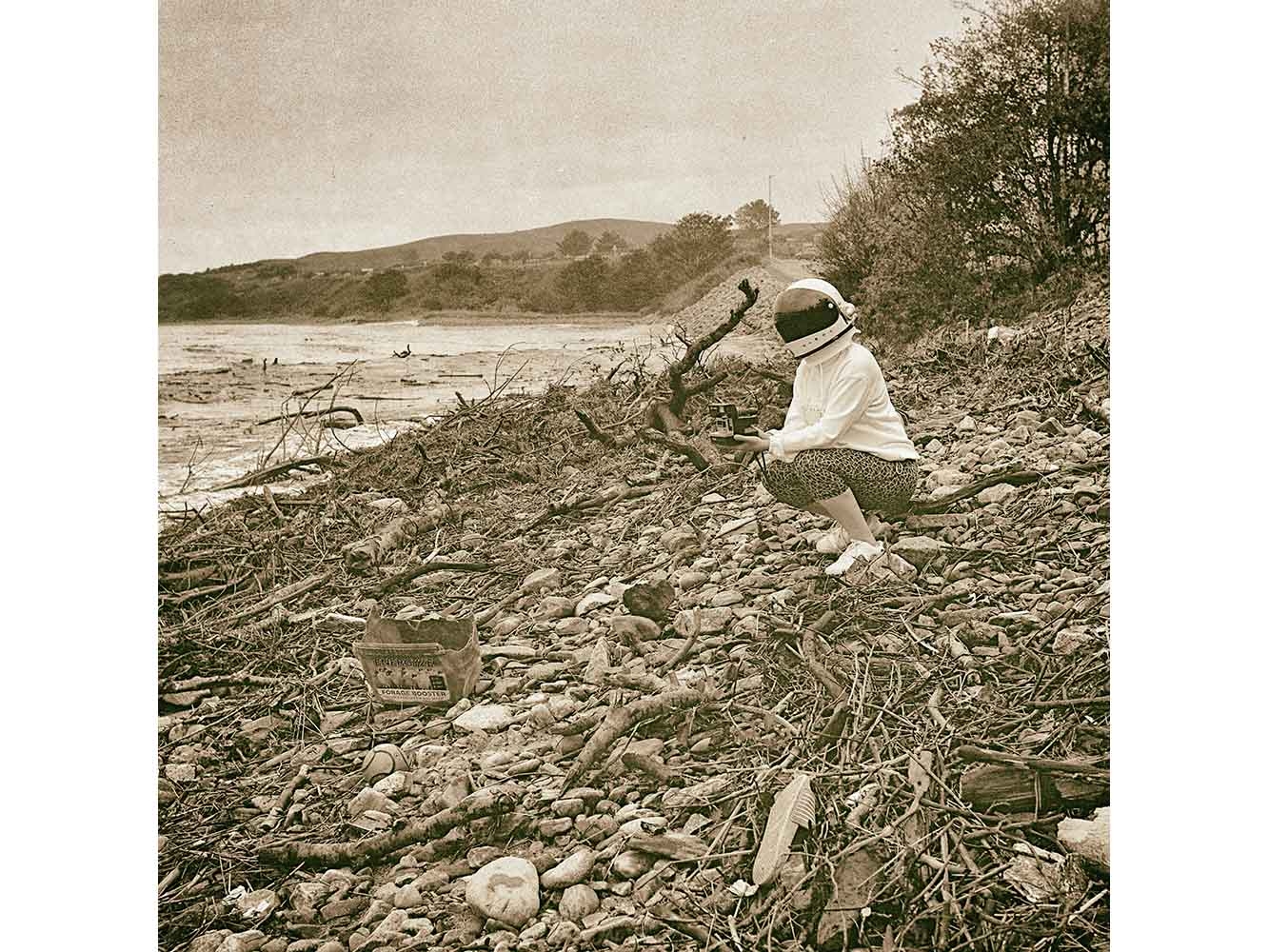 Black and white photograph of person in space helmet on a beach looking at discarded rubbish