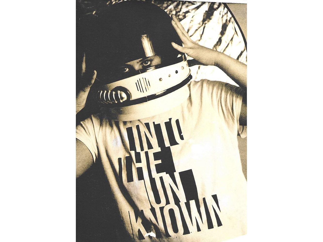Black and white photograph of person looking through visor of space helmet wearing a t-shirt that reads 'into the unknown'