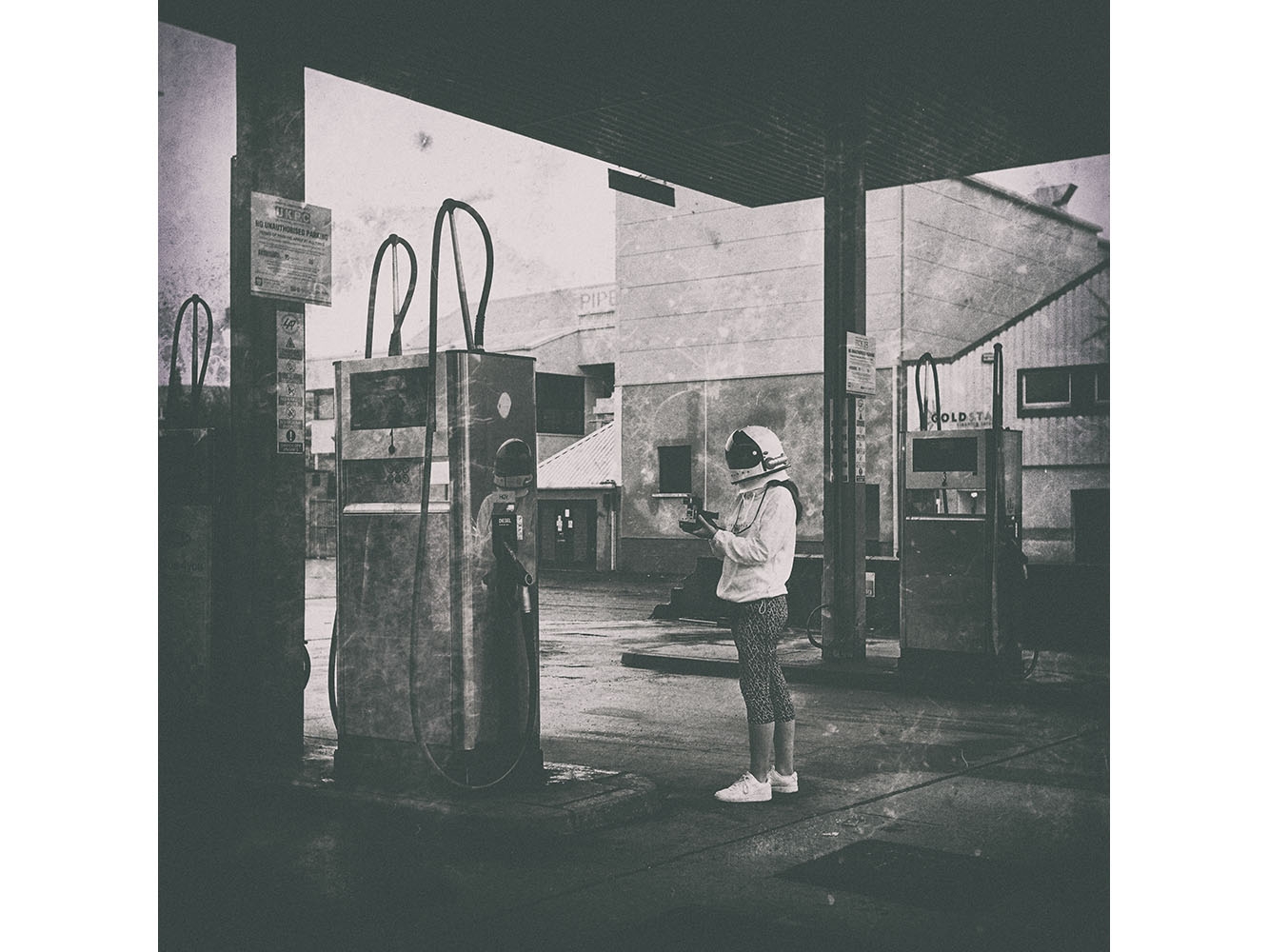 black and white photograph of person in space helmet at a petrol station