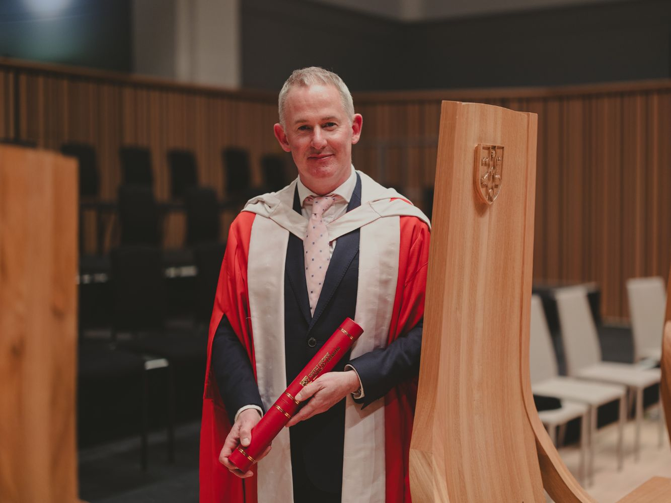 Mark standing on the stage, posing with his degree and leaning on a chair