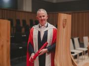 Mark standing on the stage, posing with his degree and leaning on a chair