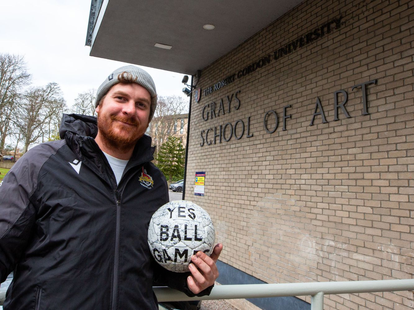 Jerome smiles, holding a football, outside Gray's School of Art on RGU campus