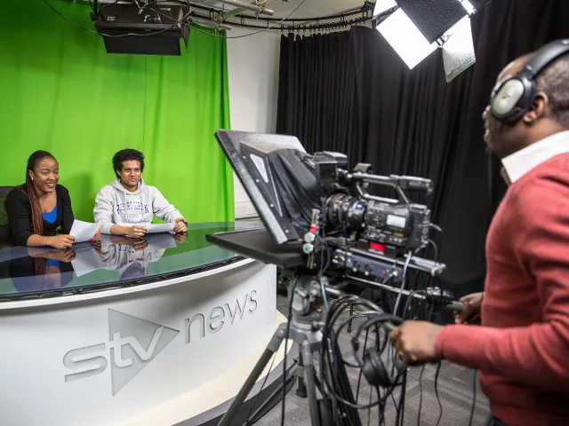 two students sitting in front of a greenscreen to be filmed for a news style report