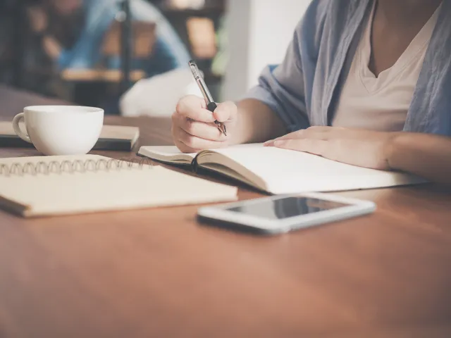 woman writing on a notebook beside coffee cup and tablet