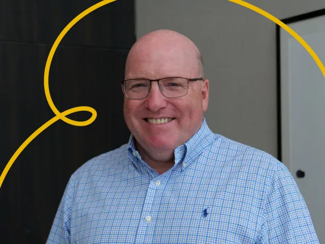 Bob Keiller smiling, wearing a blue check shirt. There is yellow UnDiscovered Aberdeen swirly branding in the background.
