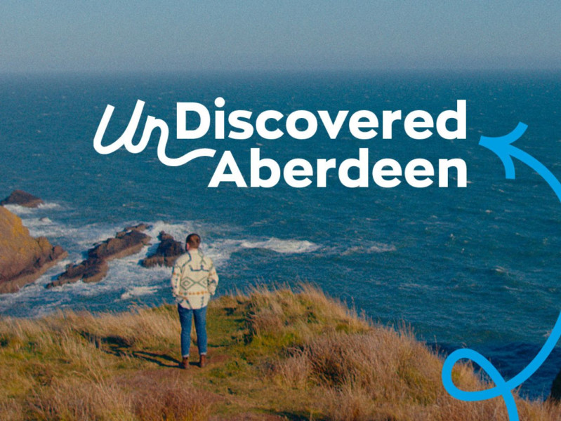 A man stands looking out from Dunnottar Castle, with UnDiscovered Aberdeen branding in the foreground
