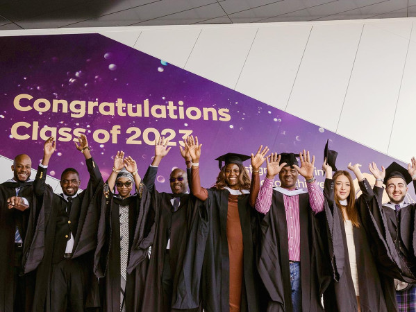 Graduates in gowns and hats with their hands in the air. Behind them is a sign saying 'Congratulations Class of 2023'