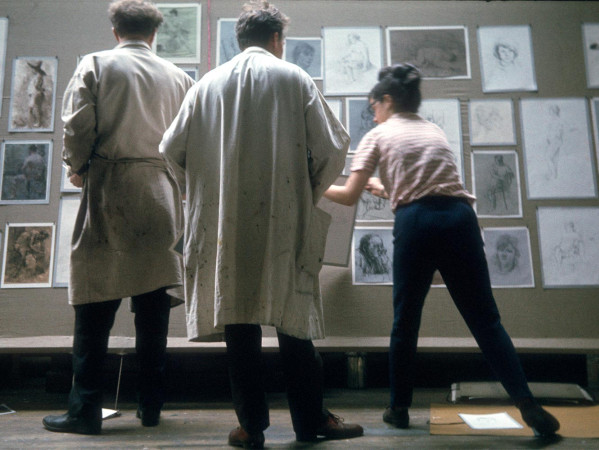 Two people wearing white lab coats and another person look at artwork on a wall
