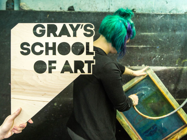 Wooden sign displaying Grays School of Art in cut out letters, held by a hand infront of someone cleaning a silkscreen