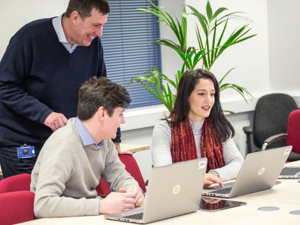 three people in an office, two sit at laptops and one stands in conversation with them